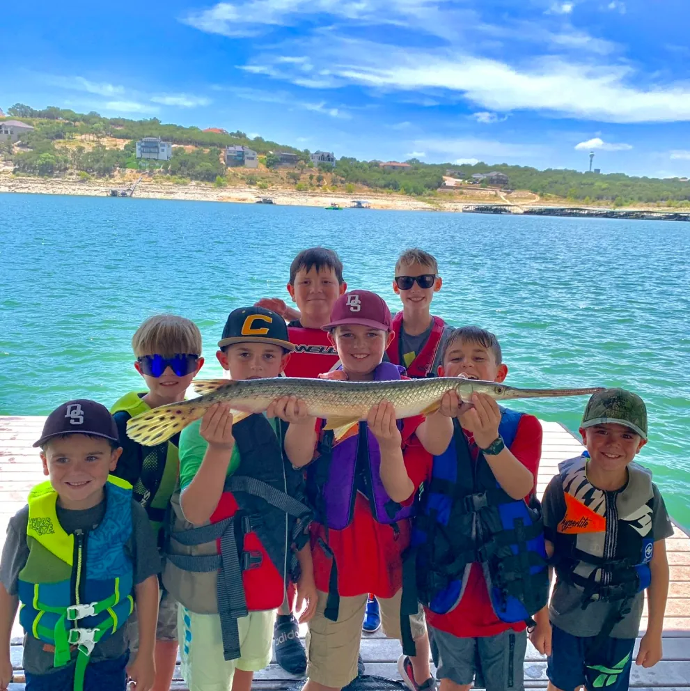 A group of young boys stand in a group while four of them work together to lift up a fish.